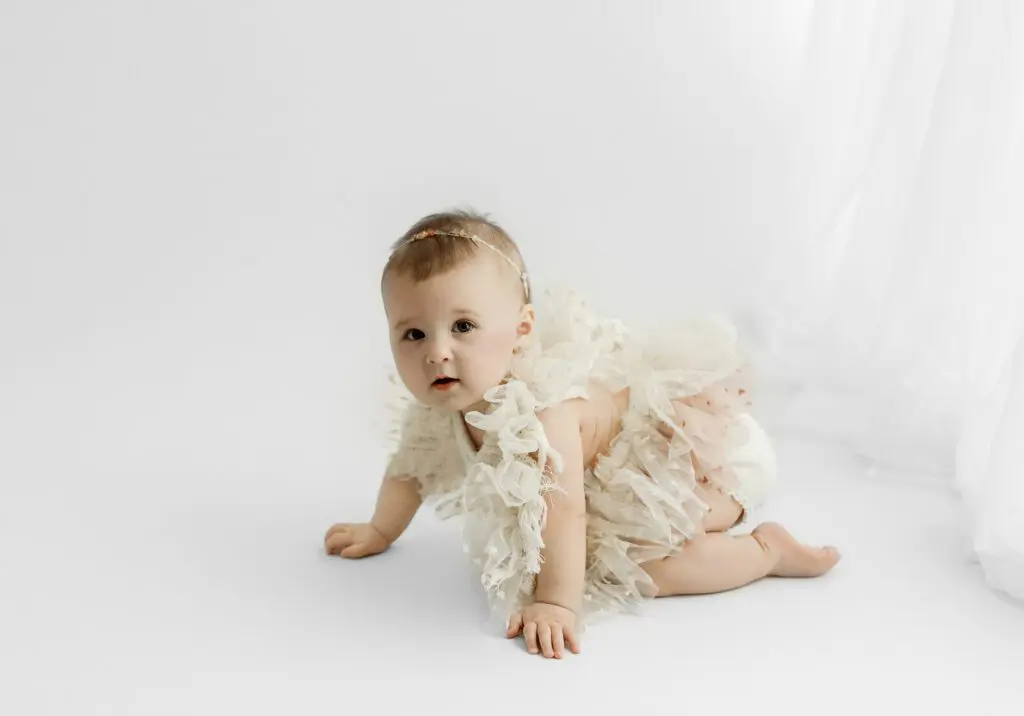 Baby Eyla crawling on a white background in a ruffled dress during her 6-month milestone photography session