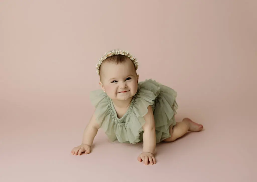 Baby Eyla crawling and smiling in a green ruffled dress with a floral headband during her 6-month milestone session on a pink background
