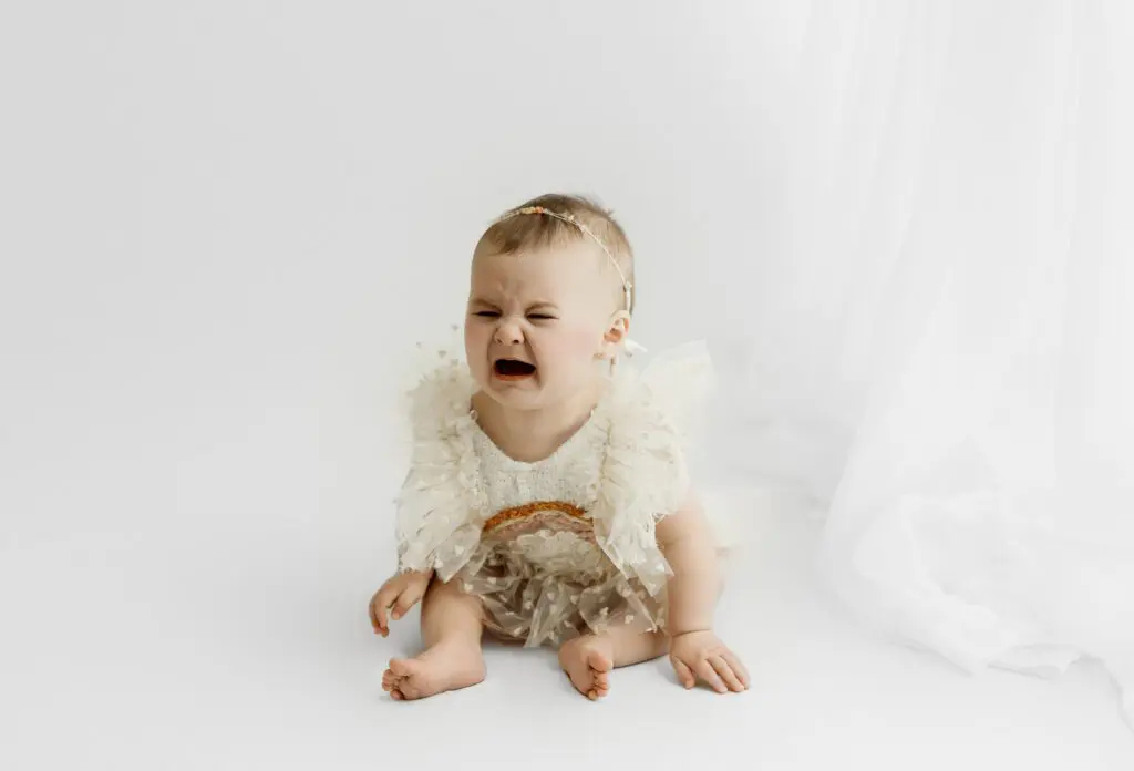 Baby Eyla crying in a ruffled dress during her 6-month milestone session on a white background