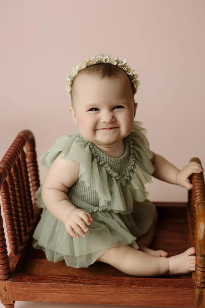 Baby Eyla sitting on a wooden bench in a green ruffled dress and floral headband, smiling during her 6-month milestone session