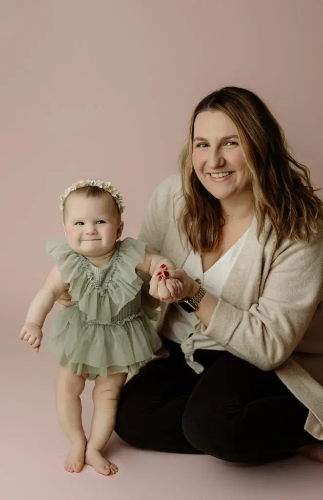 Baby Eyla holding her mom’s hand and smiling during her 6-month milestone session on a pink background