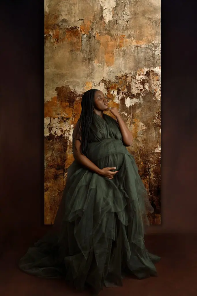 Pregnant woman in a green tulle maternity gown standing against a rustic, textured background, highlighting maternity photography in Cedar Falls, Waverly, and Waterloo, Iowa