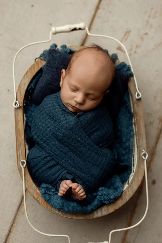 A sweet newborn baby wrapped in a blue swaddle, peacefully sleeping in a rustic basket. Captured by a professional photographer near Waterloo, Iowa, serving Cedar Falls, Waverly, and Hudson families.
