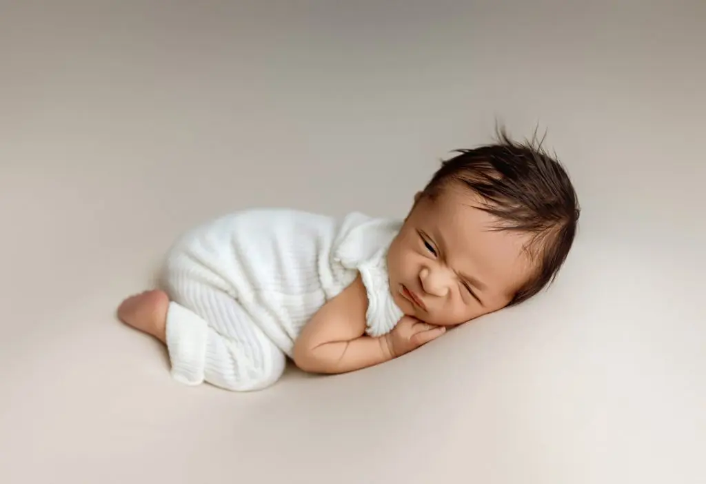 Baby Isaiah making a funny face while lying on a soft beige backdrop, dressed in a cozy white outfit during a Cedar Falls photography session