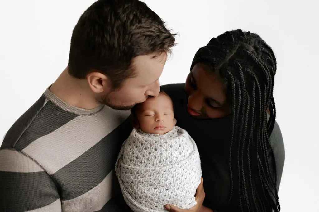 A tender moment of parents kissing their newborn baby Isaiah, wrapped in a white knit blanket, during a Cedar Falls, Iowa photography session