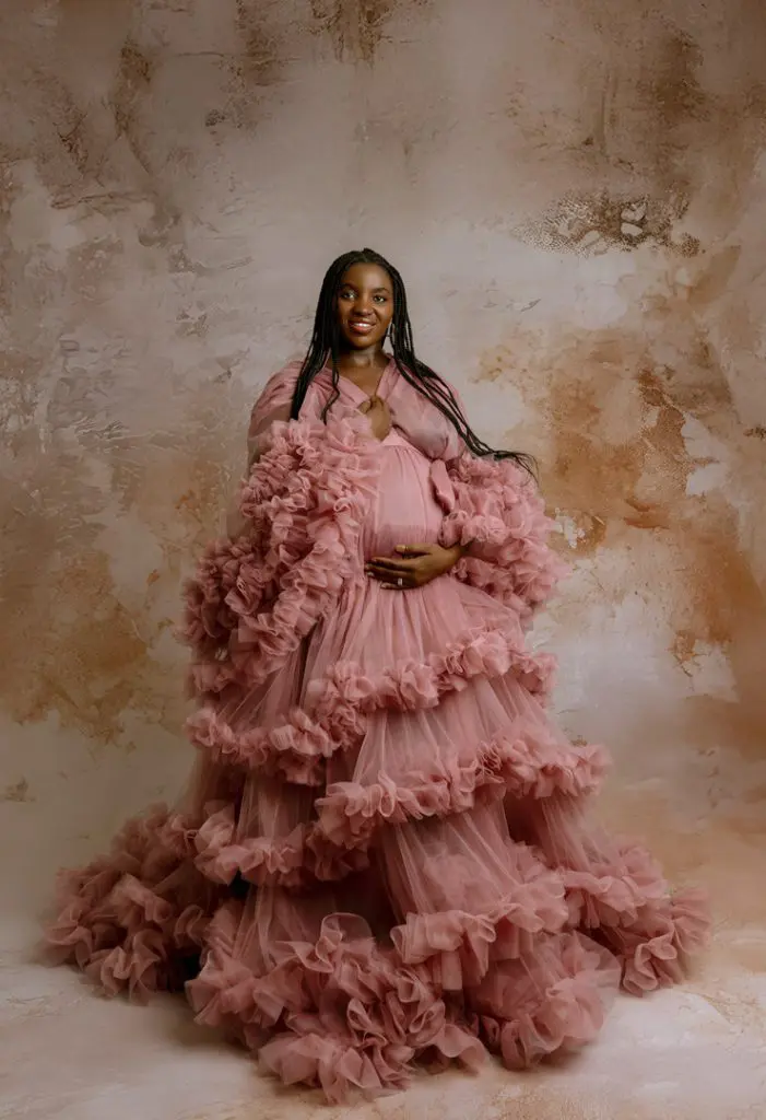 Pregnant woman in a flowing pink tulle maternity gown posing against a textured neutral background, perfect for maternity photography in Cedar Falls, Waterloo, and Waverly, Iowa