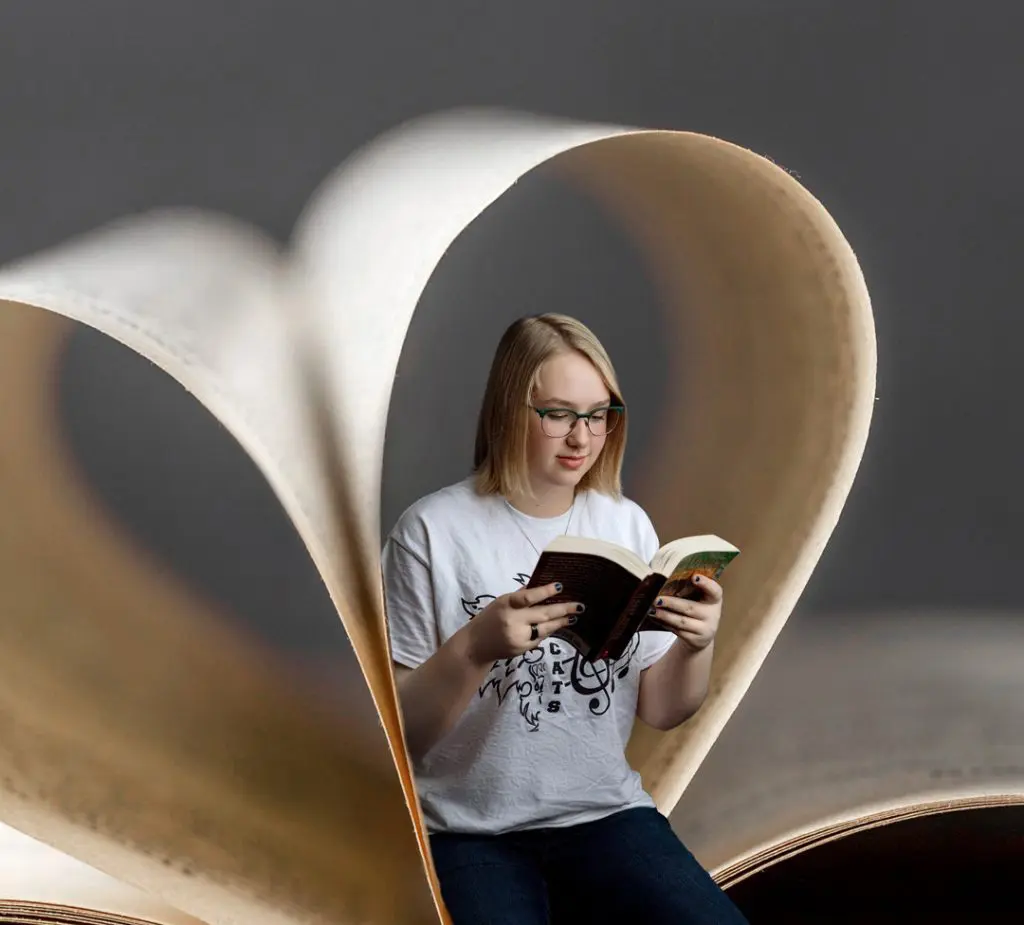 A creative portrait of a high school senior reading a book, framed by the pages of an open book folded into a heart shape. The background is a soft gray, creating a whimsical and artistic atmosphere in waverly iowa.