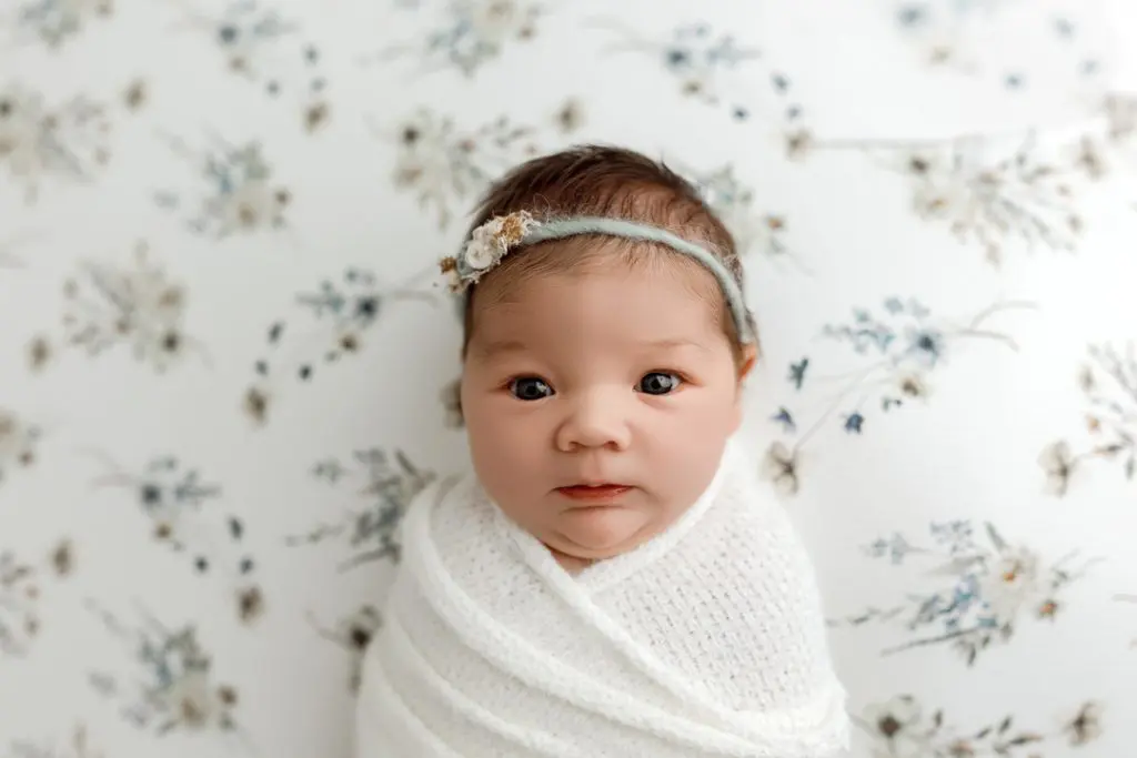 Awake newborn swaddled in white against floral backdrop during Cedar Falls photography session.