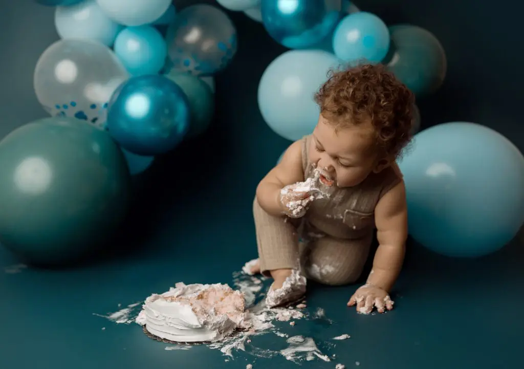 One-year-old baby enjoying a cake smash surrounded by blue balloon decorations during a milestone photography session in Cedar Falls, Iowa.