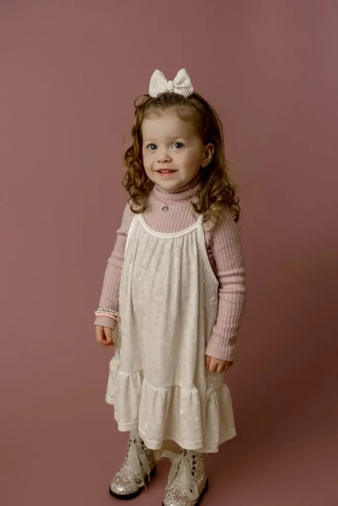 Mabel, the cutest baby contest winner, smiling in a pink sweater and white dress with sparkly boots during her mini session in Cedar Falls, Iowa.