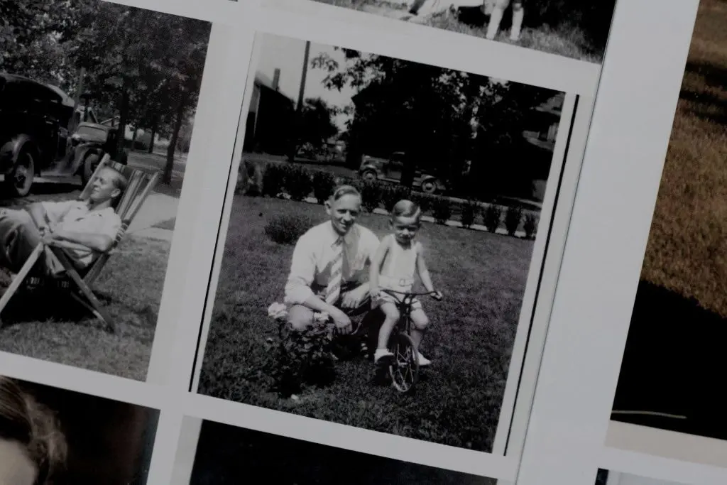 Black and white photo of a father kneeling next to child on tricycle in the yard.