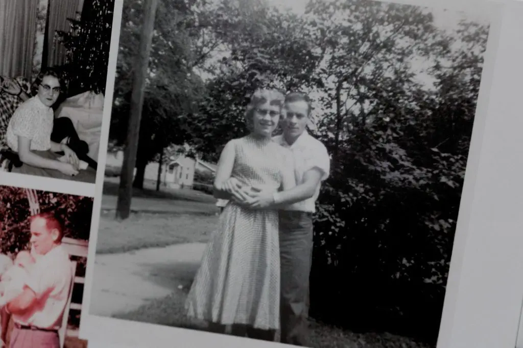 Black and white photo of grandparents hugging in a backyard, surrounded by trees.