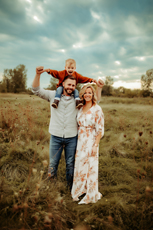 Smiling family in a grassy prairie with their son on his dad's shoulders in Cedar Falls, Iowa.