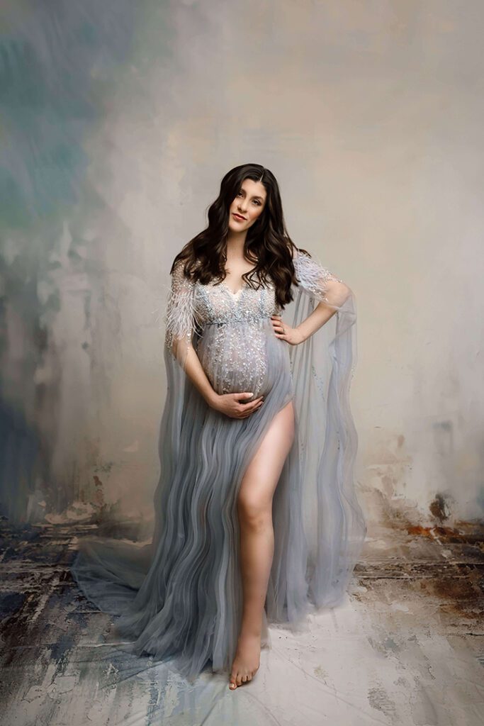A glamorous studio maternity portrait of a woman in a flowing, sheer blue gown adorned with sparkling embellishments, cradling her baby bump while standing against a textured, artistic backdrop. Perfect for showcasing elegant maternity photography in Waterloo, Iowa