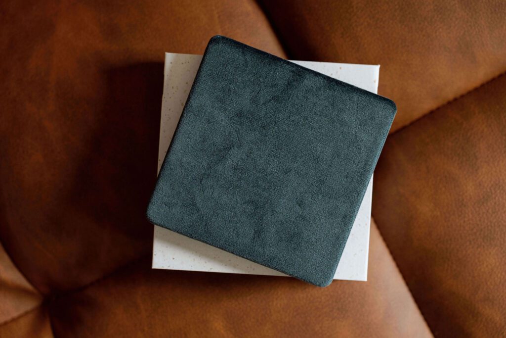 Velvet photo box in a deep teal color, perfect for storing keepsake prints, displayed on a leather couch in Cedar Falls, Iowa. Available for families in Cedar Falls, Hudson, Waverly, and Jesup.