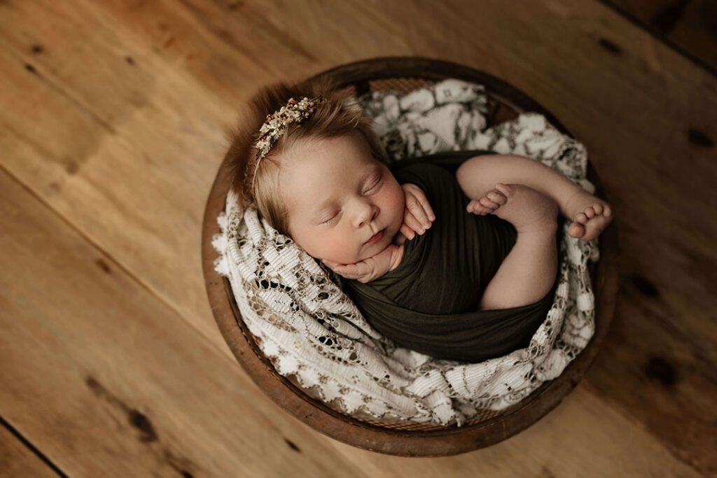 A peaceful newborn baby wrapped in a dark green swaddle, sleeping in a wooden bowl on a rustic wooden floor. Perfect for newborn photography in Cedar Falls, Iowa.