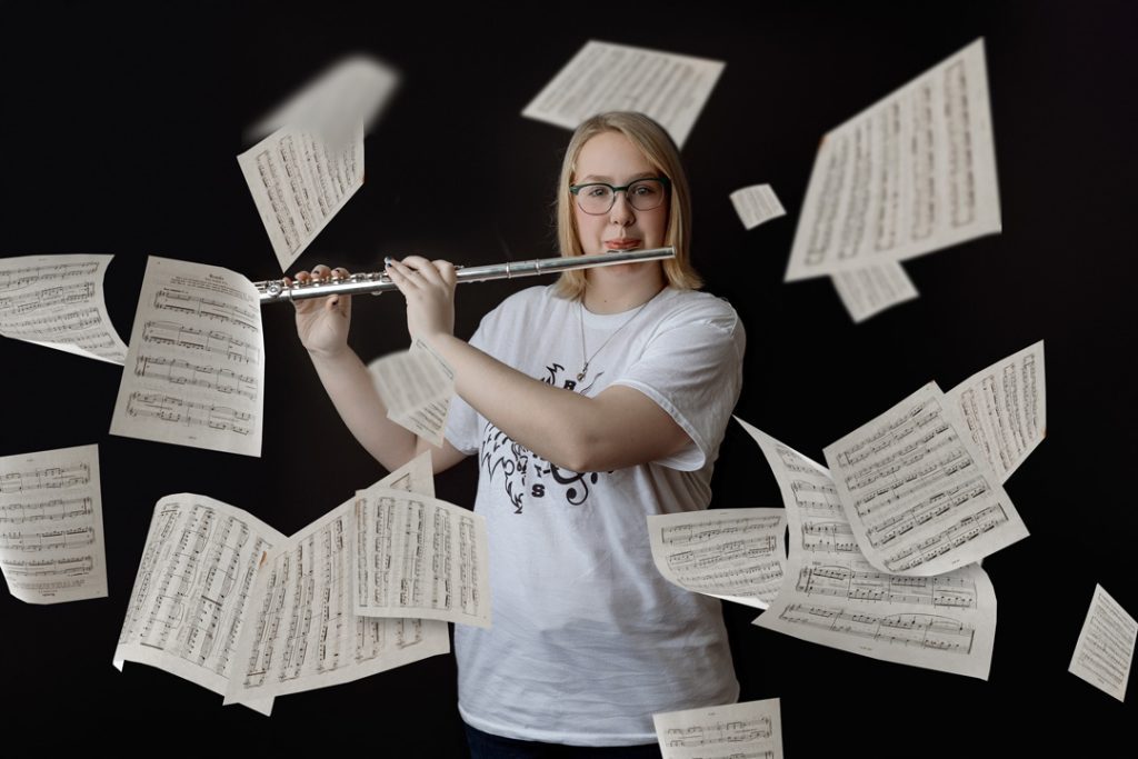 A dynamic portrait of a young woman playing the flute, surrounded by floating sheets of music. The black background highlights the movement and creativity of the scene in waterloo, iowa.