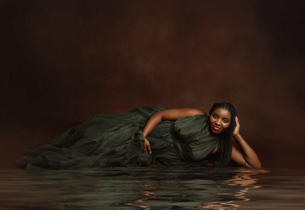 Pregnant woman in a green tulle gown lying gracefully with her reflection in water, creating a serene maternity photo for clients in Cedar Falls, Waverly, and surrounding Iowa cities