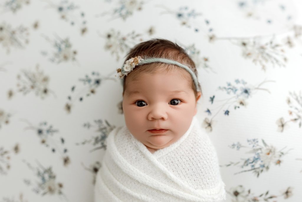 Awake newborn swaddled in white against floral backdrop during Cedar Falls photography session.