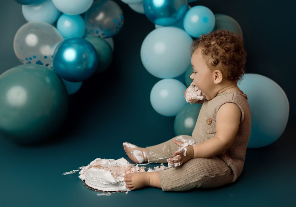 One-year-old baby enjoying a cake smash surrounded by blue balloon decorations during a milestone photography session in Cedar Falls, Iowa.