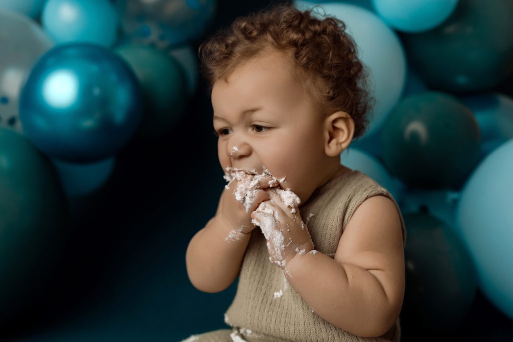 Close-up of a baby eating cake during a cake smash photography session near Waterloo, Iowa, with blue balloons in the background.