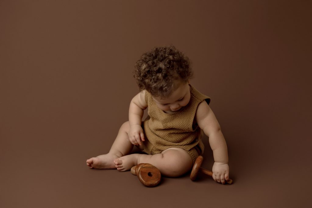 Adorable baby playing with a wooden rattle on a neutral backdrop during a newborn photography session near Waterloo, Iowa.