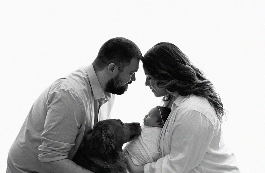Black and white portrait of parents and dog lovingly embracing newborn baby in Dike, Iowa studio.