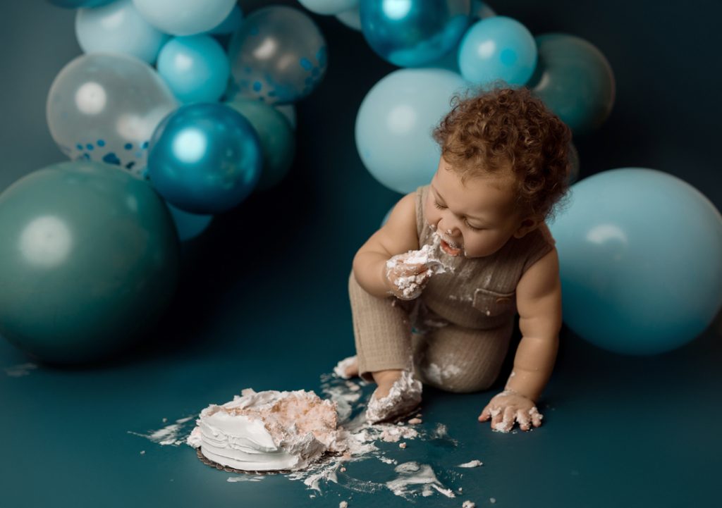 One-year-old baby enjoying a cake smash surrounded by blue balloon decorations during a milestone photography session in Cedar Falls, Iowa.