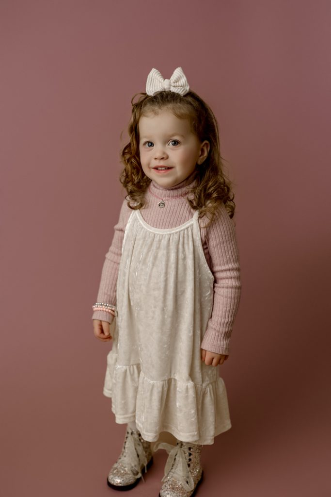 Mabel, the cutest baby contest winner, smiling in a pink sweater and white dress with sparkly boots during her mini session in Cedar Falls, Iowa.