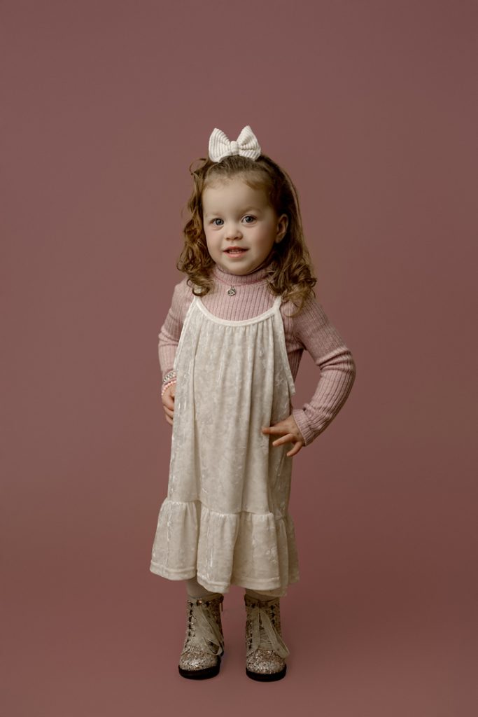 Mabel smiling in a pink sweater and white dress during her mini session as the cutest baby contest winner