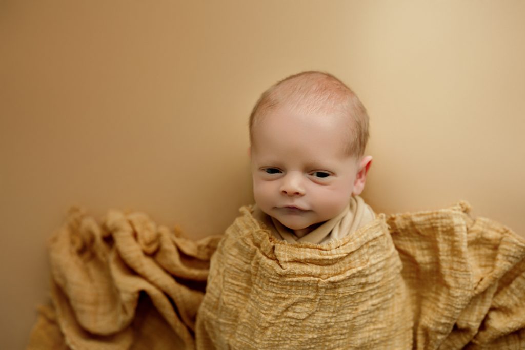 Newborn baby Hudson wrapped in a yellow blanket and looking at the camera.