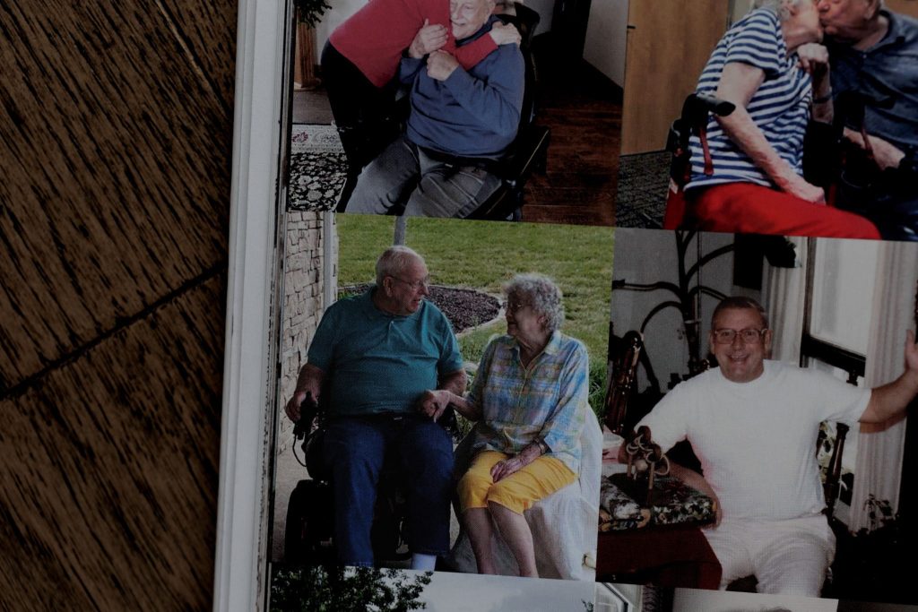 Elderly couple holding hands and smiling on a front porch with wheelchair nearby.
