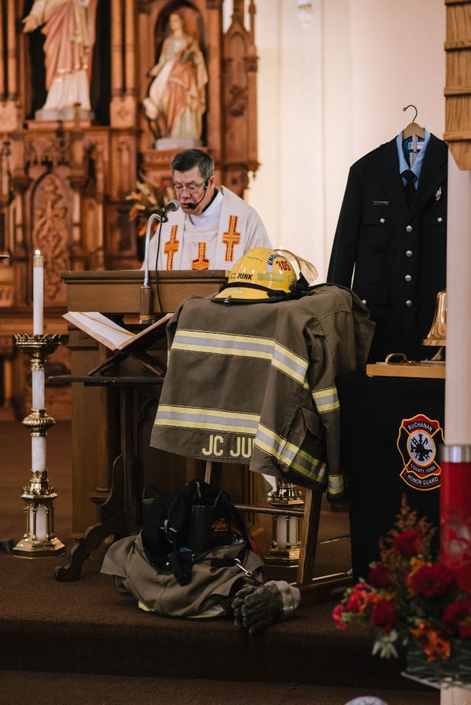 Firefighter memorial at church altar with priest reading and gear labeled JC Junk on display.