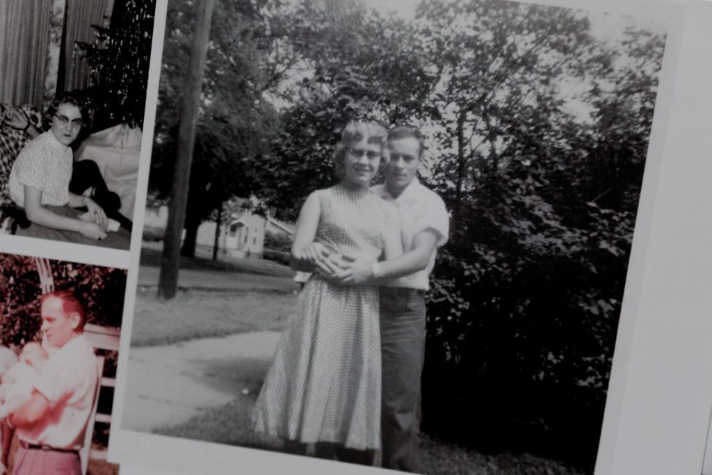 Black and white photo of grandparents hugging in a backyard, surrounded by trees.