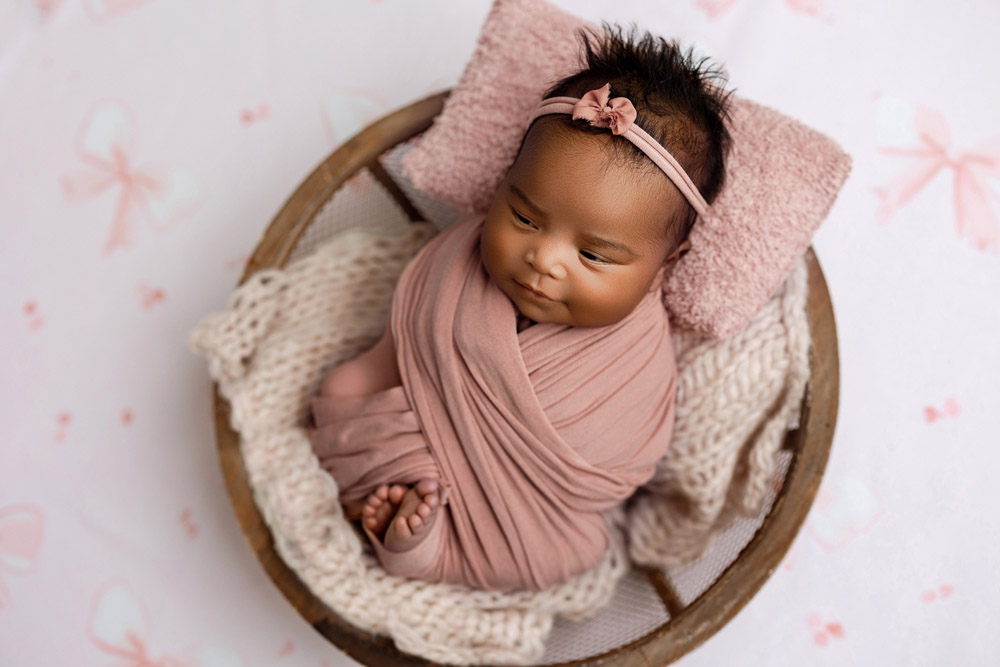 Baby Reina wrapped in dusty rose fabric, posed in a wooden bowl with pink bows backdrop in Waverly, Iowa