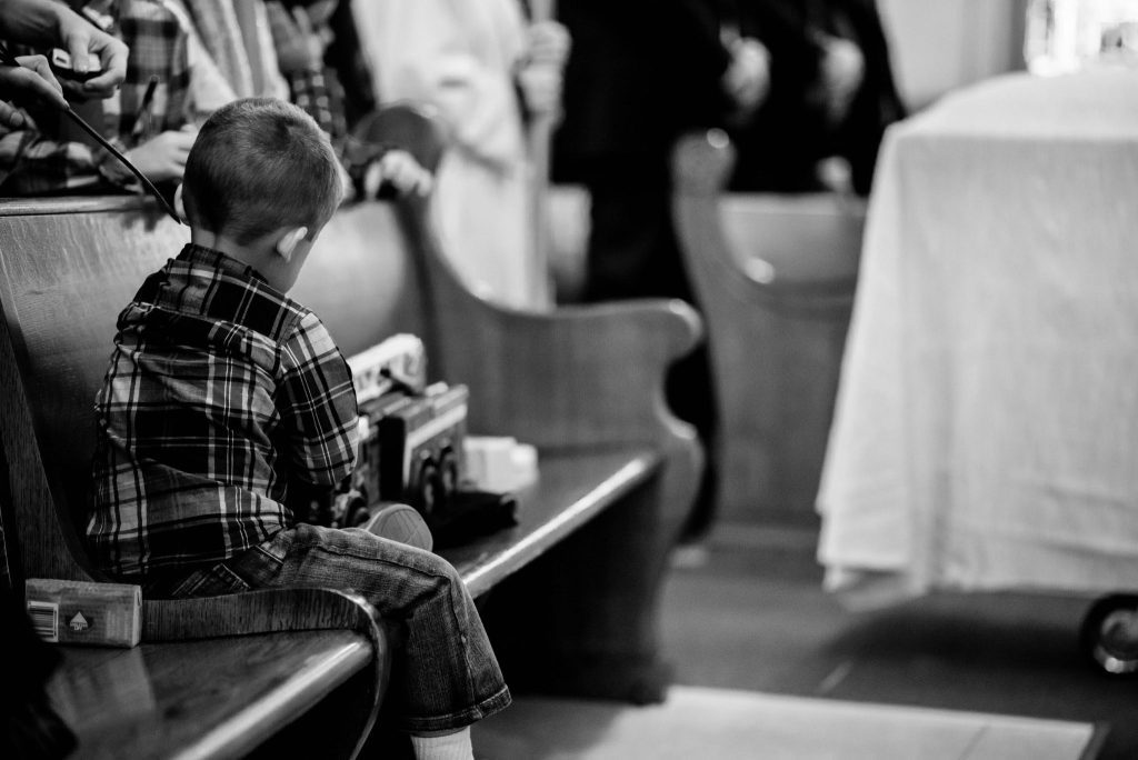 Black and white image of a young boy sitting on a pew at a funeral, playing quietly with toy fire trucks.